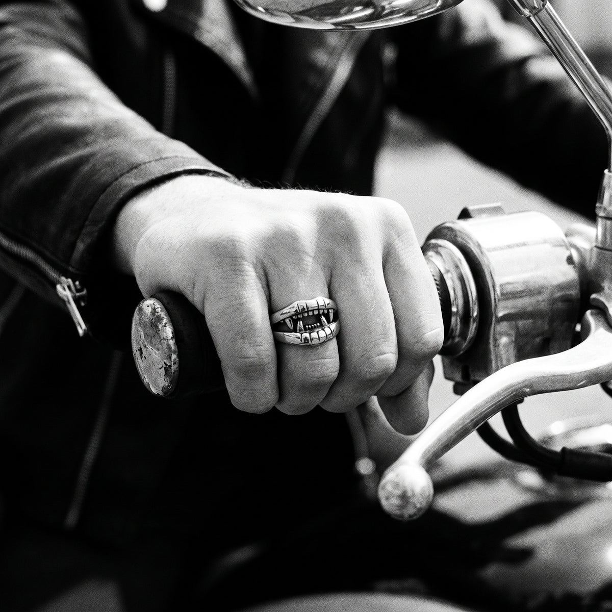 Young man's hand in a black leather jacket gripping a motorcycle handlebar, wearing the Fang devil mouth ring, black and white 35mm film