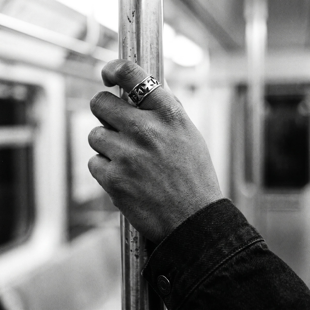 Man's hand gripping a metal subway pole wearing the Sacrament cross band ring, black and white film photo