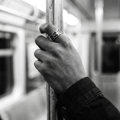 Man's hand gripping a metal subway pole wearing the Sacrament cross band ring, black and white film photo