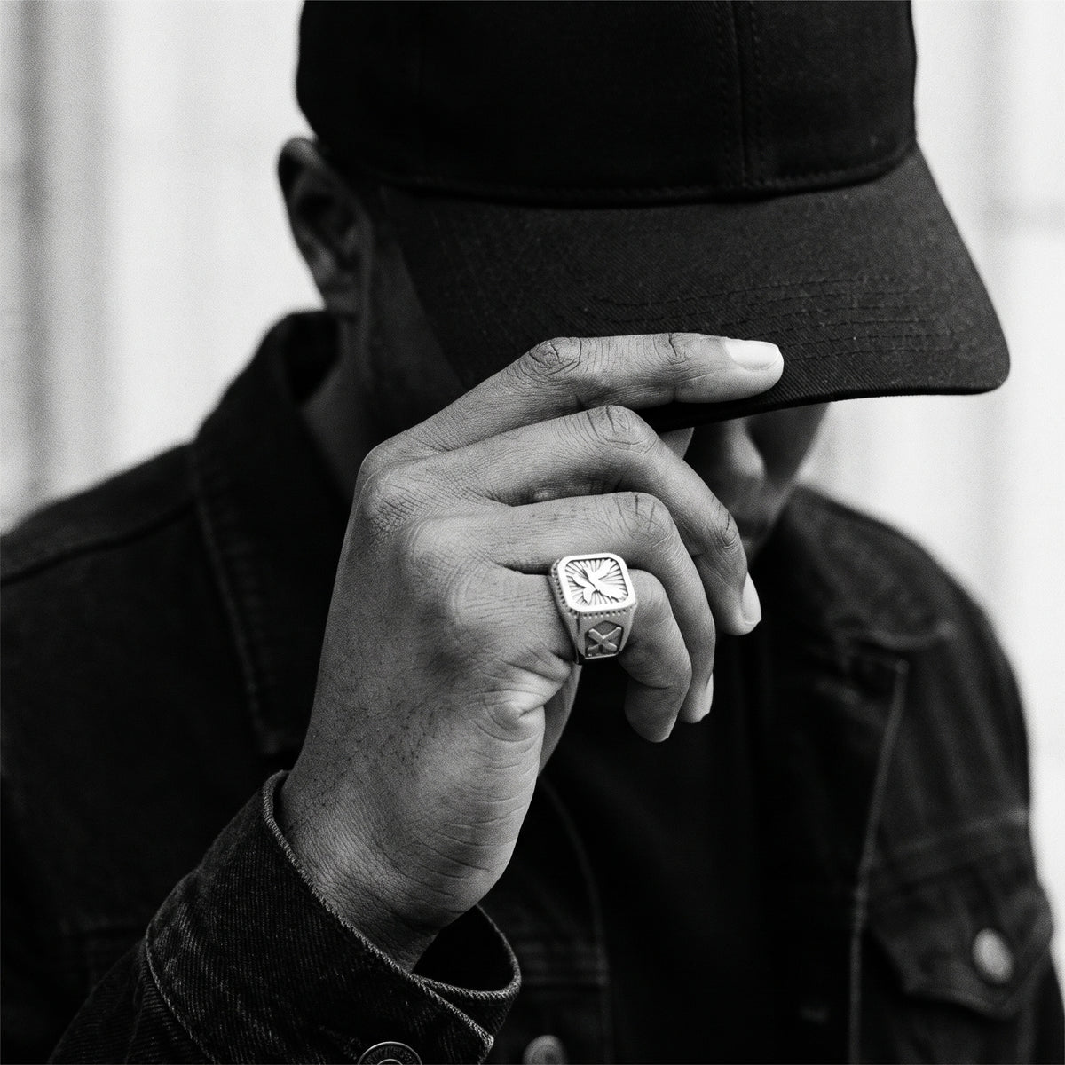Black and white photo of a hand lifting the brim of a black cap wearing the Talon winged eagle signet ring