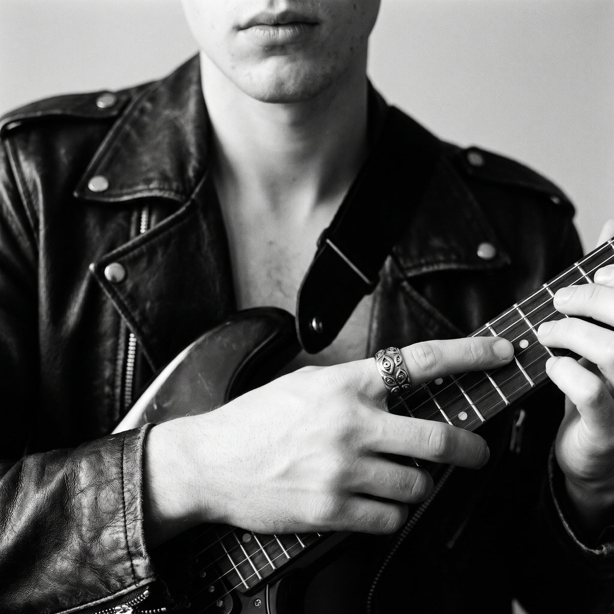 Young man's hand on a black electric guitar neck, in a black leather jacket, wearing the Watcher eye ring, black and white 35mm film