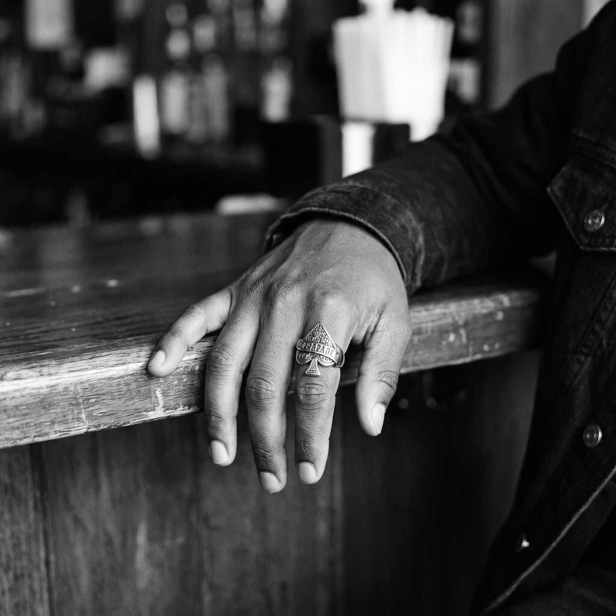 Man's hand resting on a wooden bar counter wearing the Wildcard ace of spades ring, black and white film photo